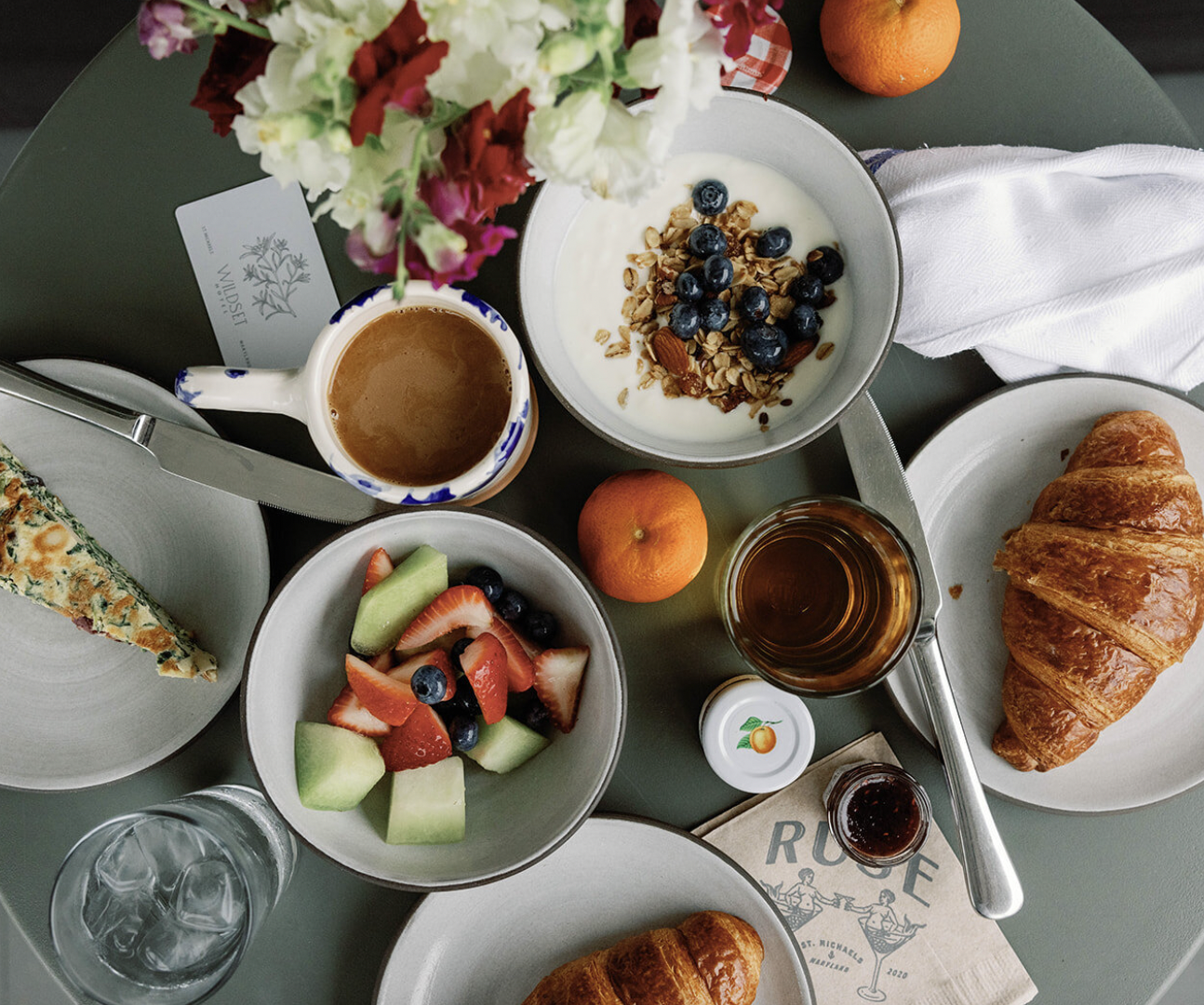Overhead view of a breakfast table with a croissant, fresh fruit salad, yogurt with granola and blueberries, a slice of spinach and tomato quiche, a cup of coffee, a glass of water, a small jar of jelly, a bowl of orange segments, and an orange. Ther