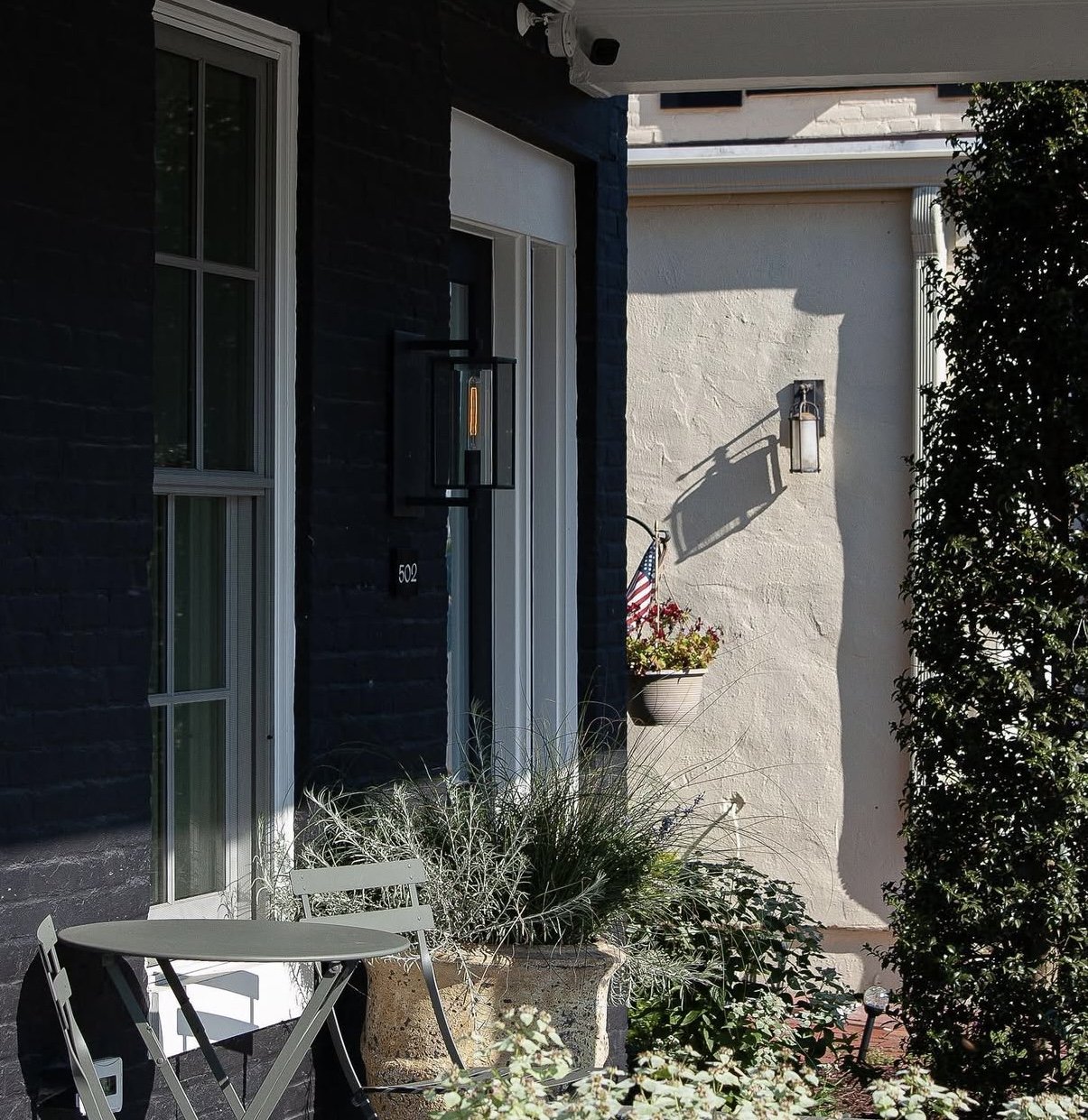 Front porch of a house with a black brick wall, white door, and outdoor lighting fixtures. There are potted plants, a small table with two chairs, and an American flag in a pot. Shadows of the fixtures are cast on the wall.