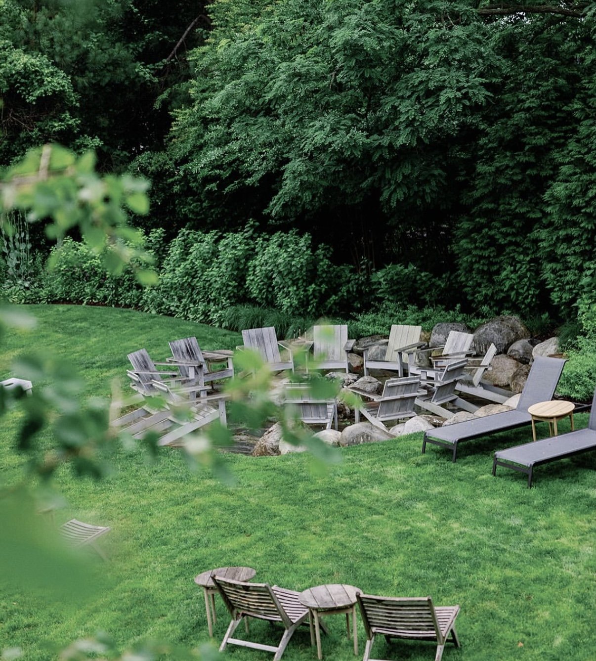 A backyard with lush green grass, surrounded by dense trees and bushes. There are outdoor lounge chairs and Adirondack chairs arranged around a stone fire pit, with some stacking rocks nearby.
