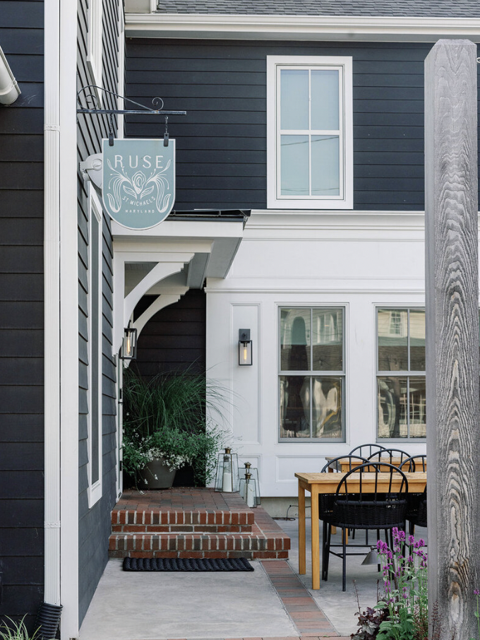 Outdoor patio area with black and white house exterior, brick steps, potted plants, outdoor table with black chairs, and a sign reading 'RUSE St. Michaels Maryland'.