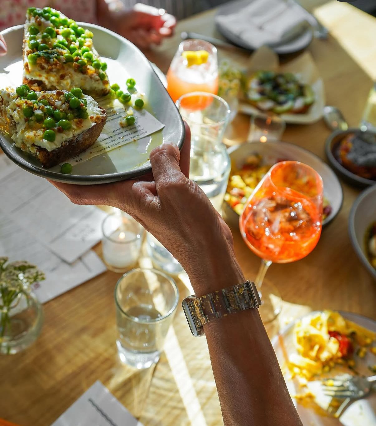 Person holding a plate with two slices of cake topped with green peas, surrounded by drinks and plates of food on a wooden table.