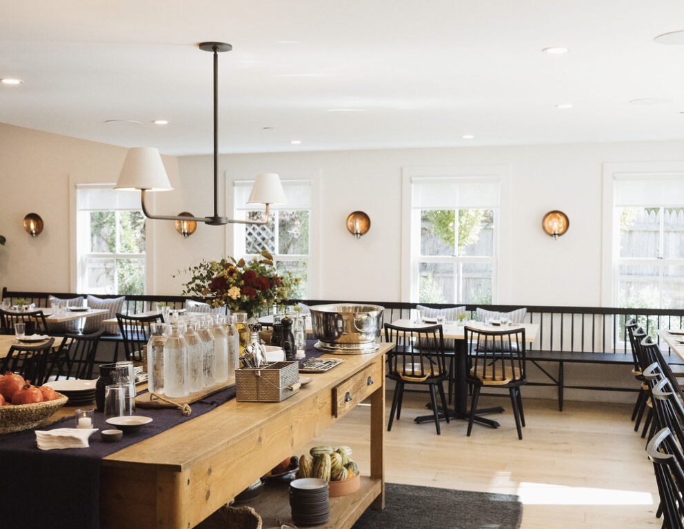 Interior of a dining room with tables, chairs, and a wooden buffet table holding glass bottles, a large metal bowl, and decorative items. Bright natural light coming through windows.