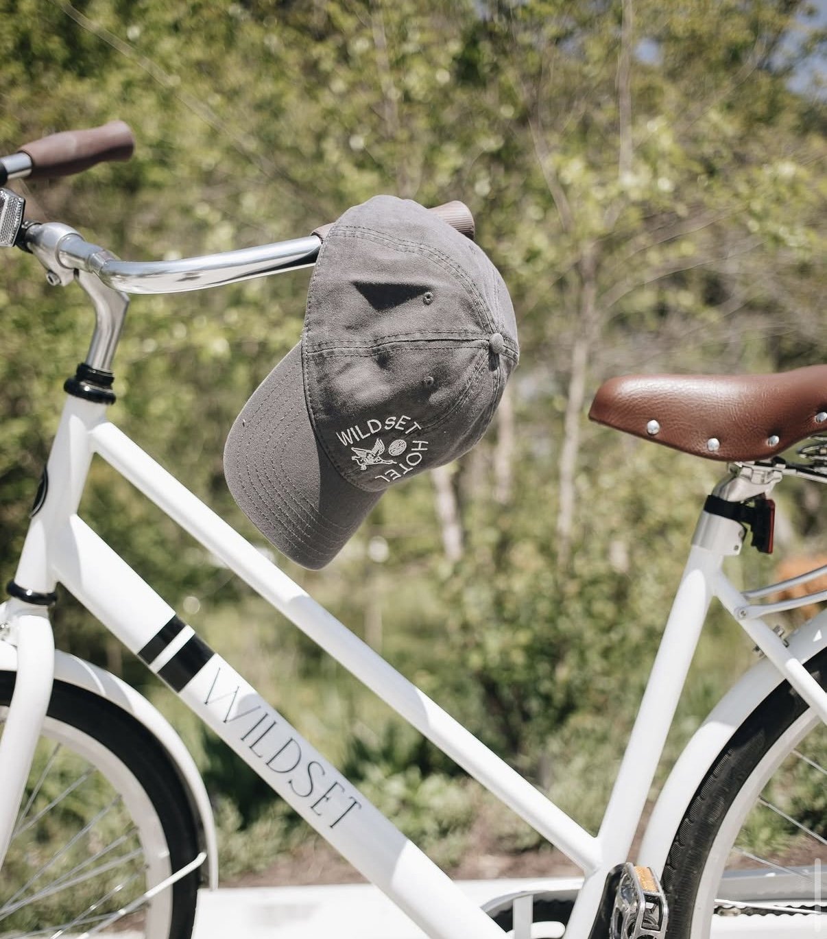 A white WILDSENT bicycle with a brown leather seat and a gray cap hanging from the handlebars, against a background of green trees.