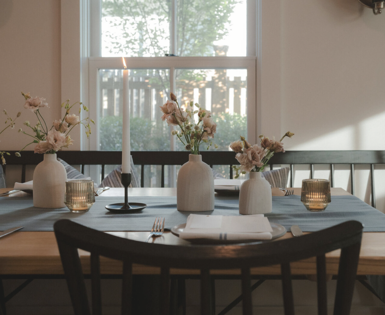A decorated dining table with three white vases holding pink flowers, a lit candle, and two amber-colored candle holders, set near a window with sunlight streaming in.