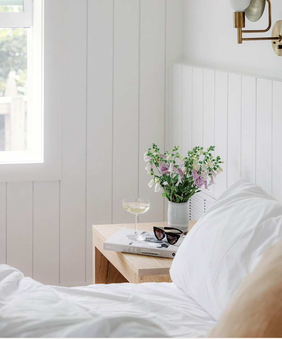 A bedroom with a white wooden-paneled wall, a window, a wooden nightstand with a vase of pink and white flowers, a glass of wine, sunglasses, and a magazine.