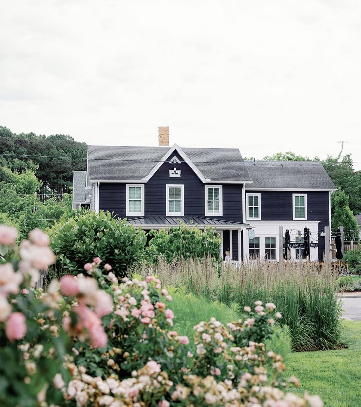 A two-story house painted dark blue with white trim, surrounded by lush green plants and pink flowers in the foreground, under a partly cloudy sky.