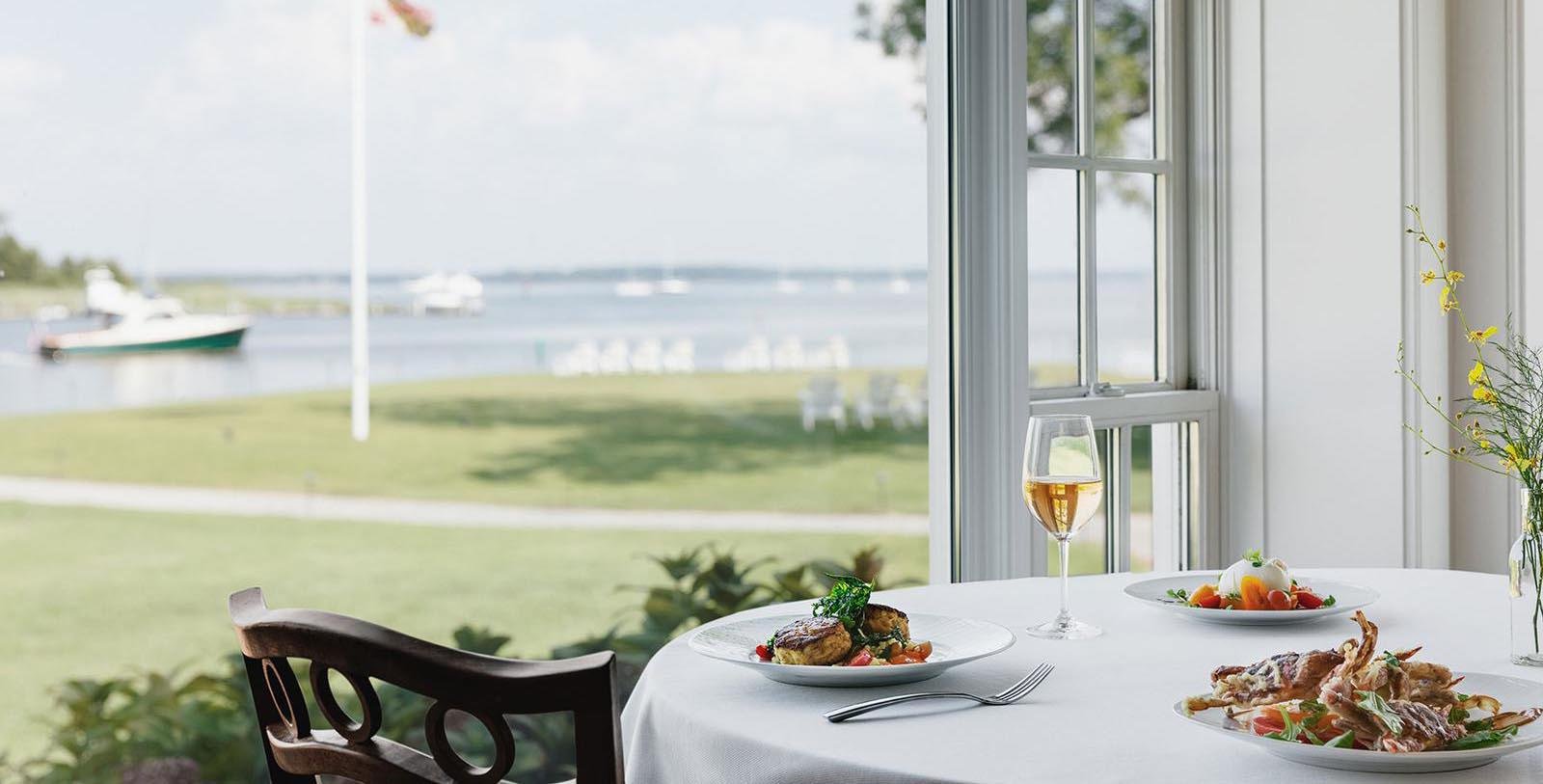 Dining table set with plates of food, a glass of white wine, and a vase with yellow flowers, overlooking a waterfront view through a window.