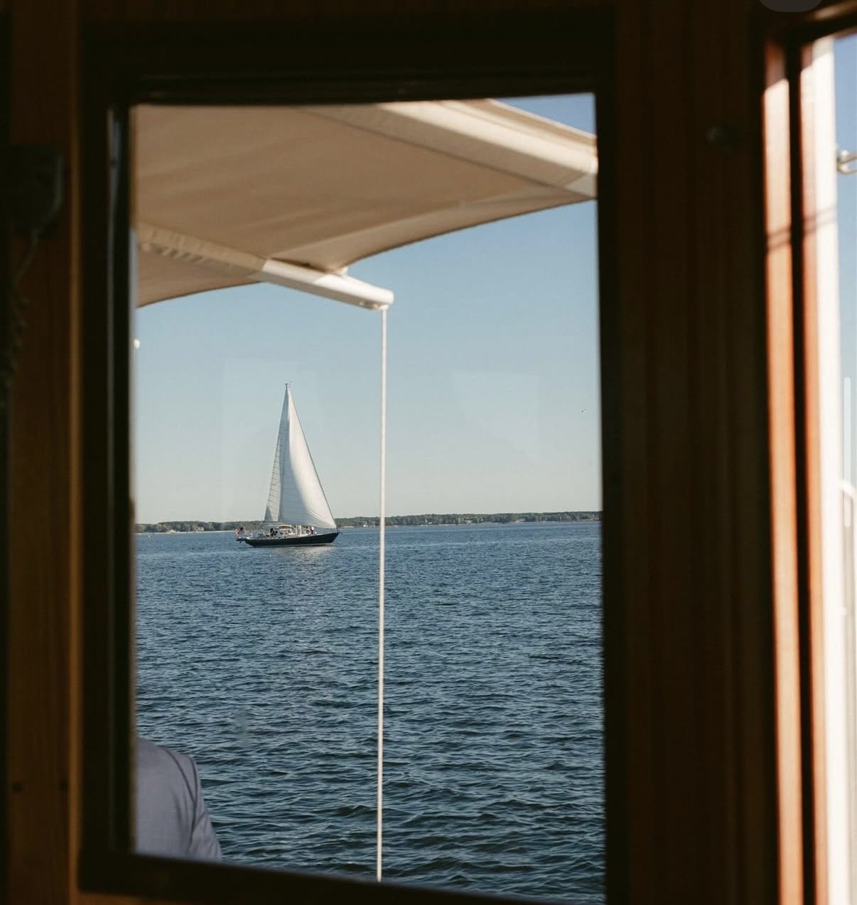 View of a sailboat on the water seen through a window with a wooden frame. The sailboat has a white sail and appears to be sailing on a calm body of water under a clear sky.
