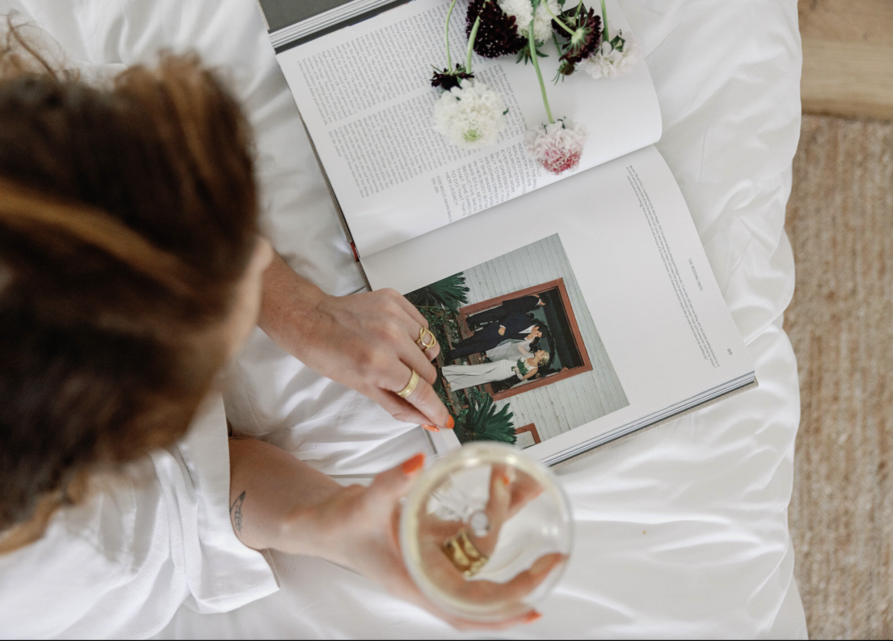 A person with a ring on their finger is sitting on a bed, holding a glass of champagne, and looking at a magazine with photos and text. There are pink and white flowers resting on the magazine.