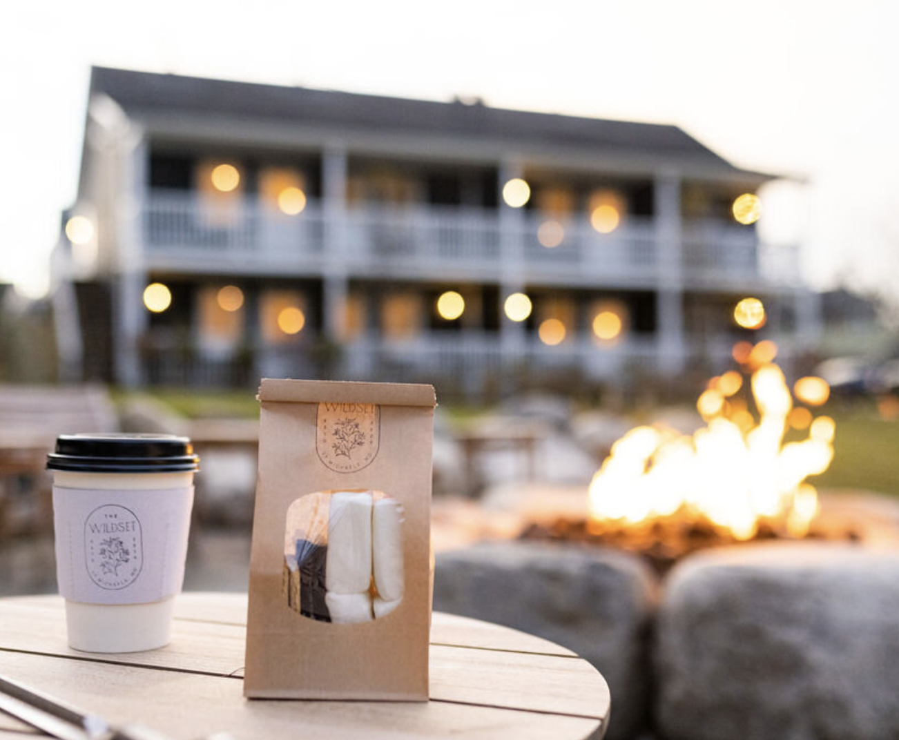 Coffee cup and marshmallows on a wooden table outdoors during evening with a house and firepit in the background, with warm lighting and bokeh effect.