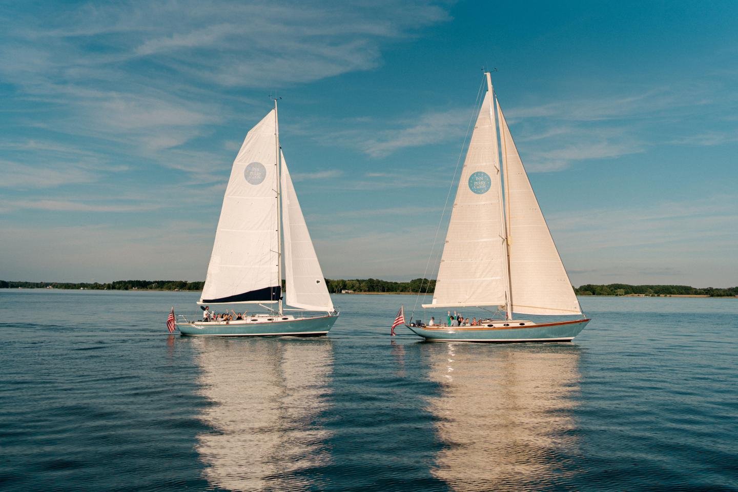 Two sailboats with white sails on calm water, both with American flags at the stern, under a blue sky with wispy clouds.