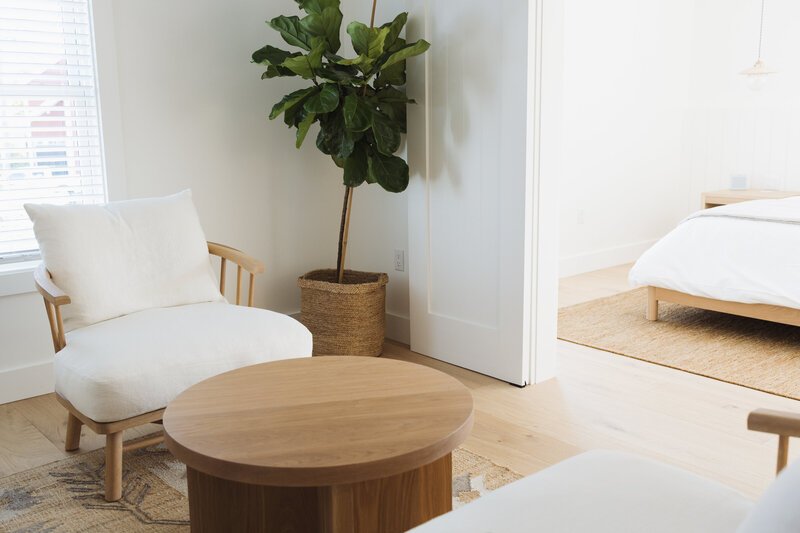 Bright bedroom with white walls, a large green potted plant, a wooden armchair with a white cushion, a round wooden coffee table, and a bed with white bedding on a light-colored wooden floor.