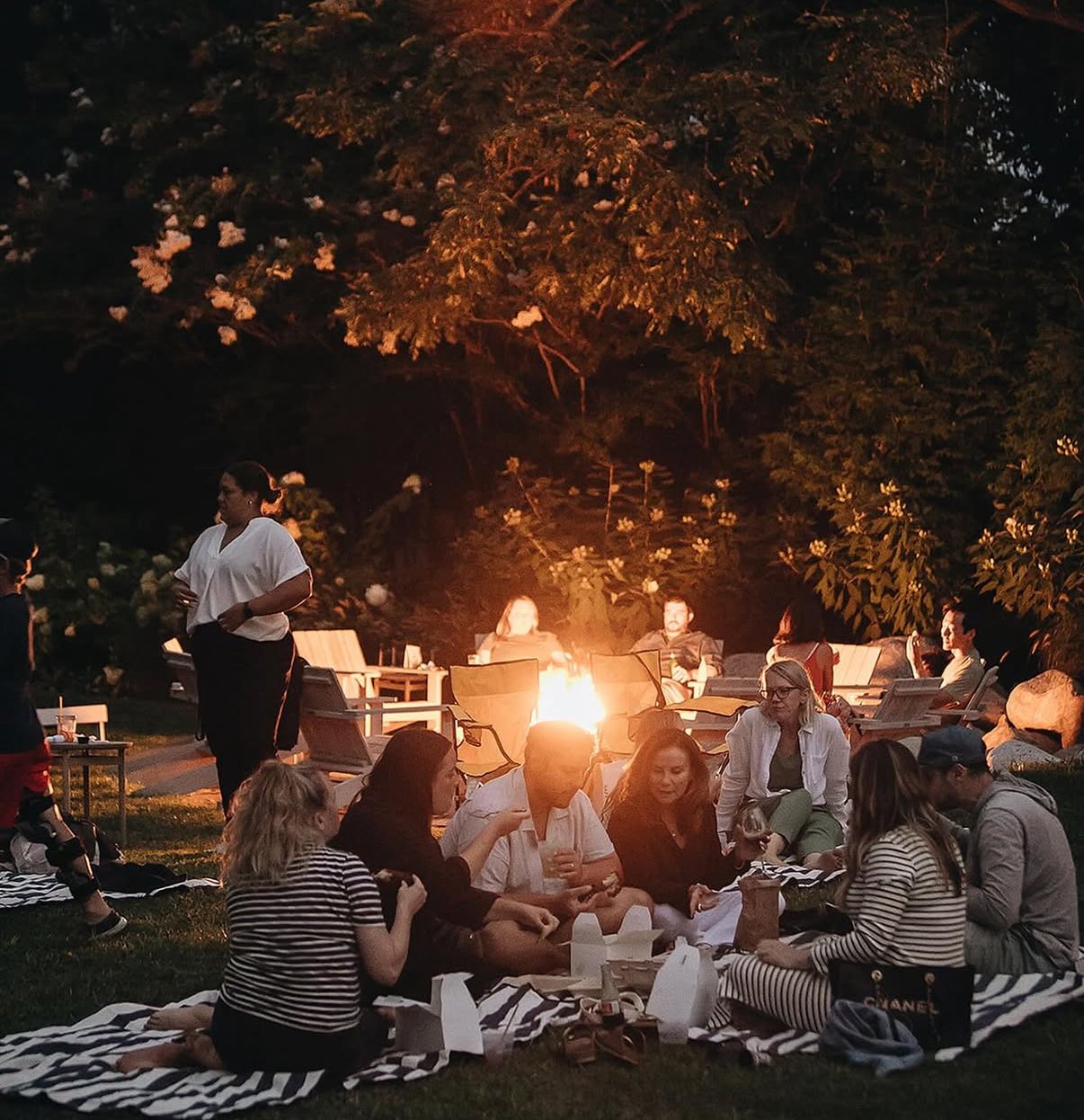 A group of people having a picnic outdoors at sunset, sitting on blankets near a fire, with trees in the background.