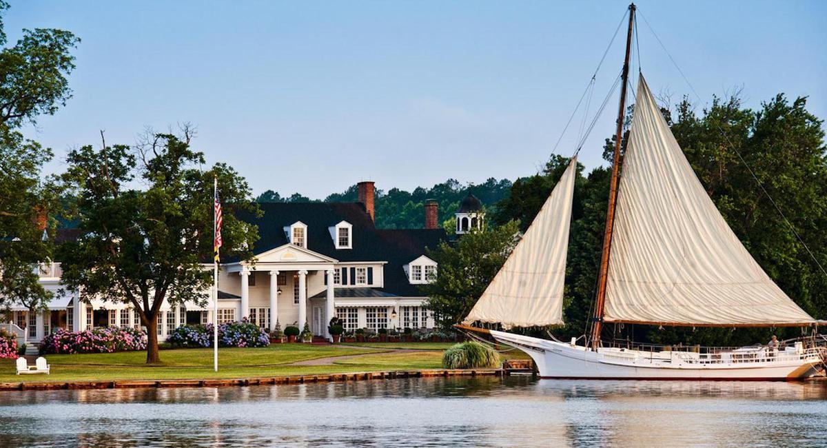 Large white house with columns, surrounded by trees and flowering bushes, next to a body of water with a sailboat docked nearby.