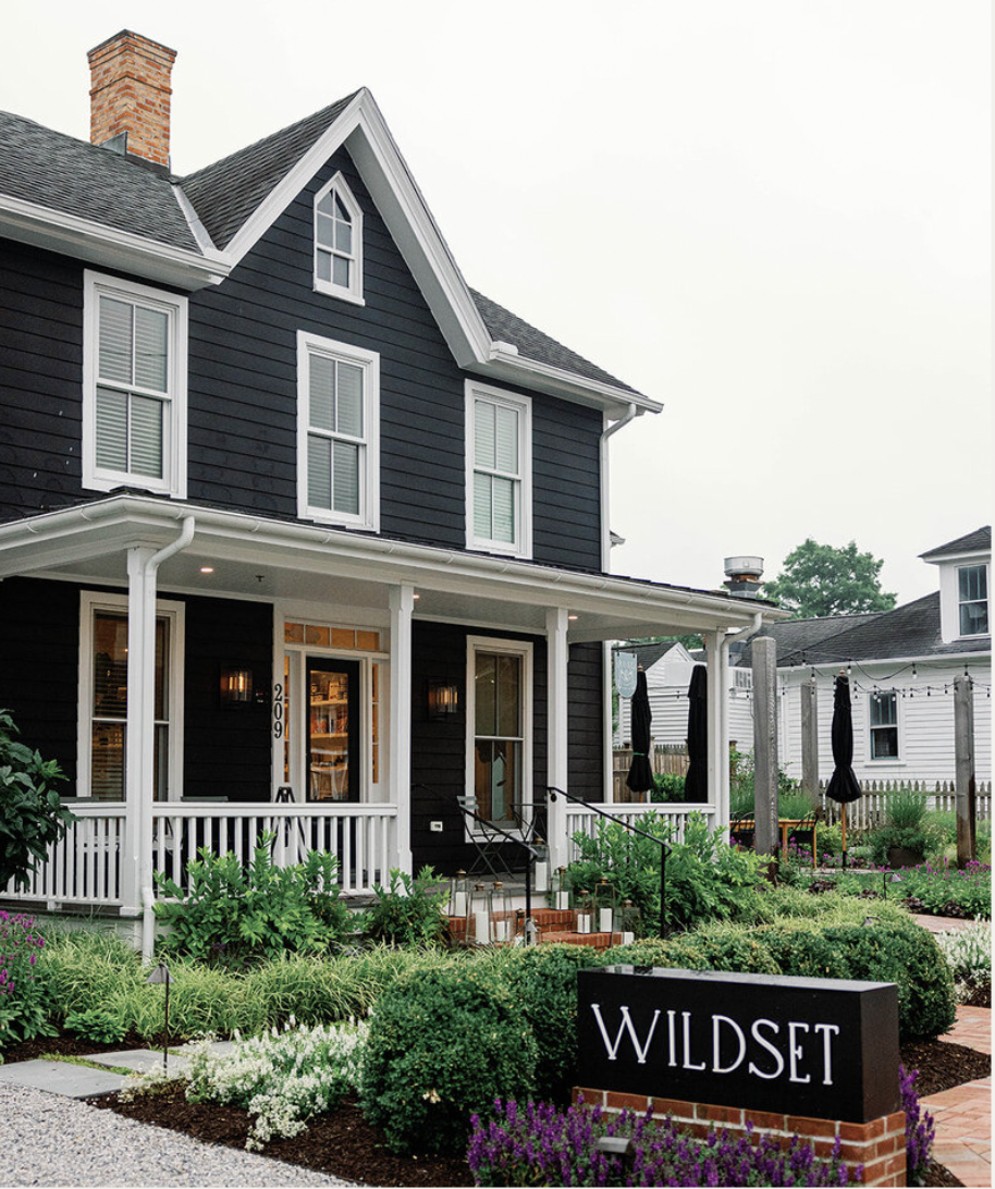 Front yard of a dark gray house with white trim, a porch with white railing, and a garden with various plants. A sign reads 'WILDS' in front of the garden.