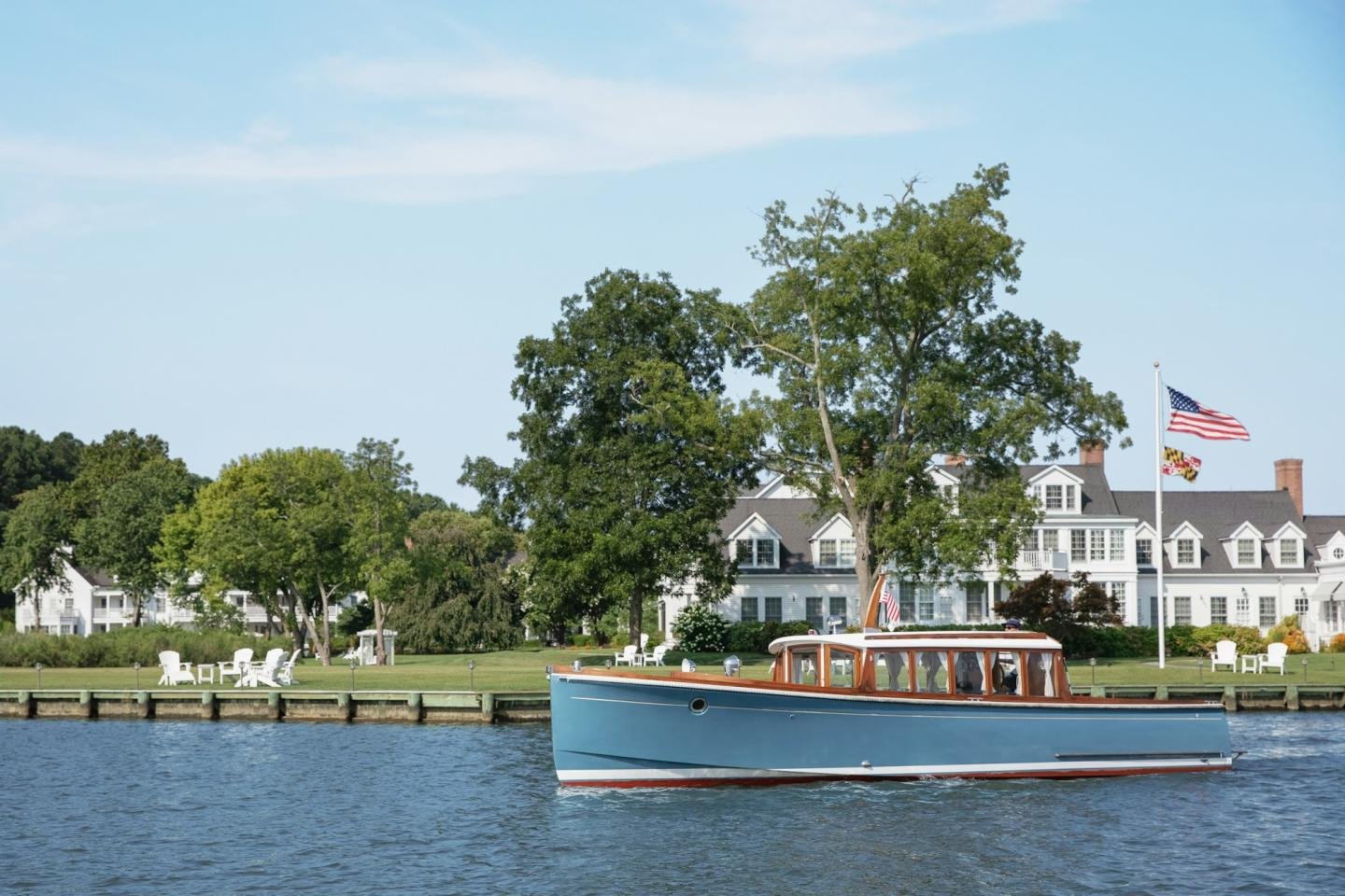 A blue boat with a wooden cabin floats on a river near a shoreline with green trees, white chairs, and a large white house, under a clear blue sky. American flags are on display.