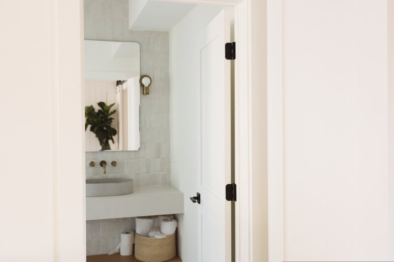 A minimalist bathroom with a wall-mounted mirror, a countertop sink, a small round light fixture, a potted plant reflection in the mirror, and a basket with rolled towels underneath.