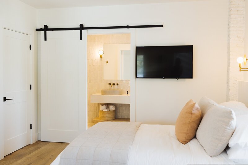 A cozy bedroom with a white bed and pillows, a mounted flat-screen TV, a sliding barn door leading to a bathroom, and light fixtures on the wall.