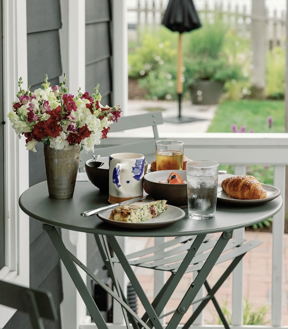 Breakfast on a small gray patio table with a vase of pink and white flowers, a slice of vegetable quiche, a croissant, a cup, a glass of tea, a glass of water, and a bowl of strawberries with blueberries. Outside view of a garden with green plants, a