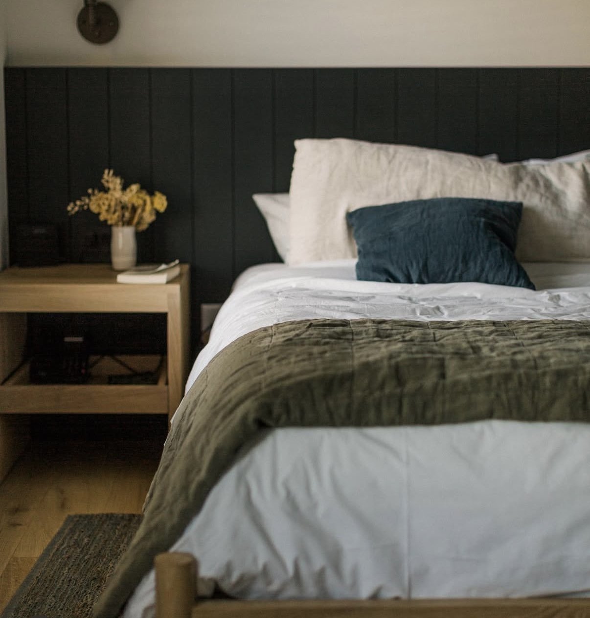 A neatly made bed with white linens, a green throw blanket, and a dark teal pillow in a bedroom. A wooden nightstand with a vase of yellow flowers, a book, and a small black box is beside the bed, with a dark-paneled wall behind.