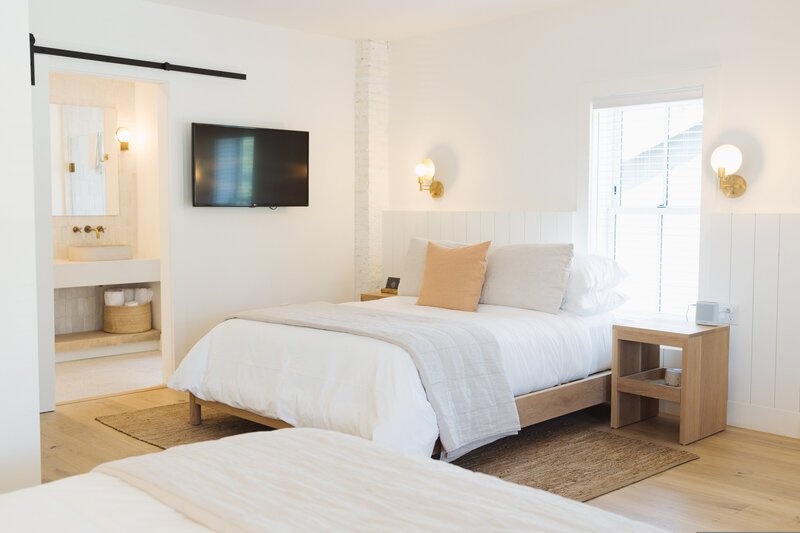 Bright bedroom with white walls and bedding, two bedside lamps, a wooden nightstand, a wall-mounted TV, and a window letting in natural light. An open doorway reveals a bathroom with a sink and mirror.