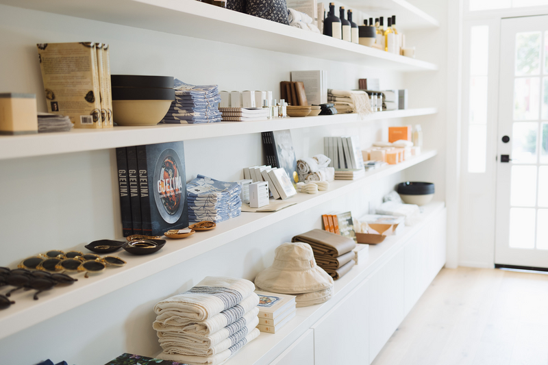 White retail display shelves filled with various home goods such as bowls, towels, books, and bottles, near a door with glass panes.