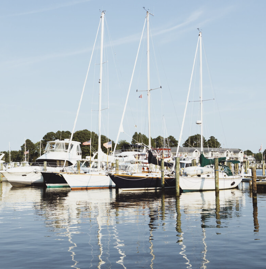 Multiple boats docked at a marina with calm water, American flags, and trees in the background under a partly cloudy sky.