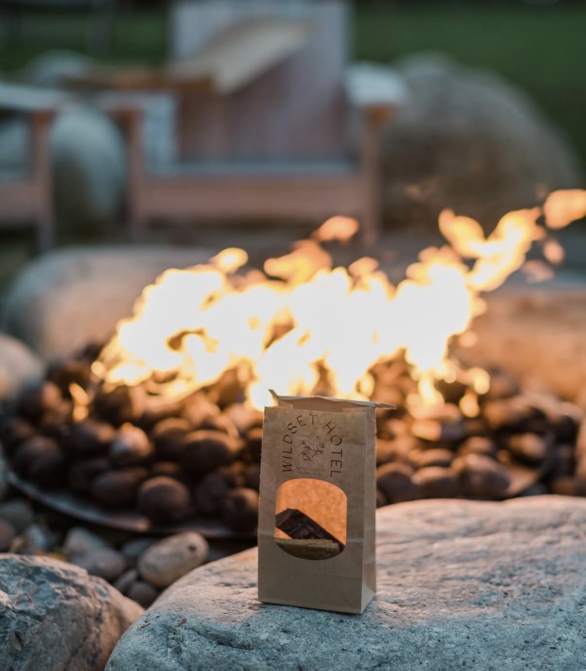 A small paper bag with a cut-out window displaying a sunny landscape logo, placed on a rock in front of a campfire surrounded by stones, with a blurred outdoor seating area in the background.