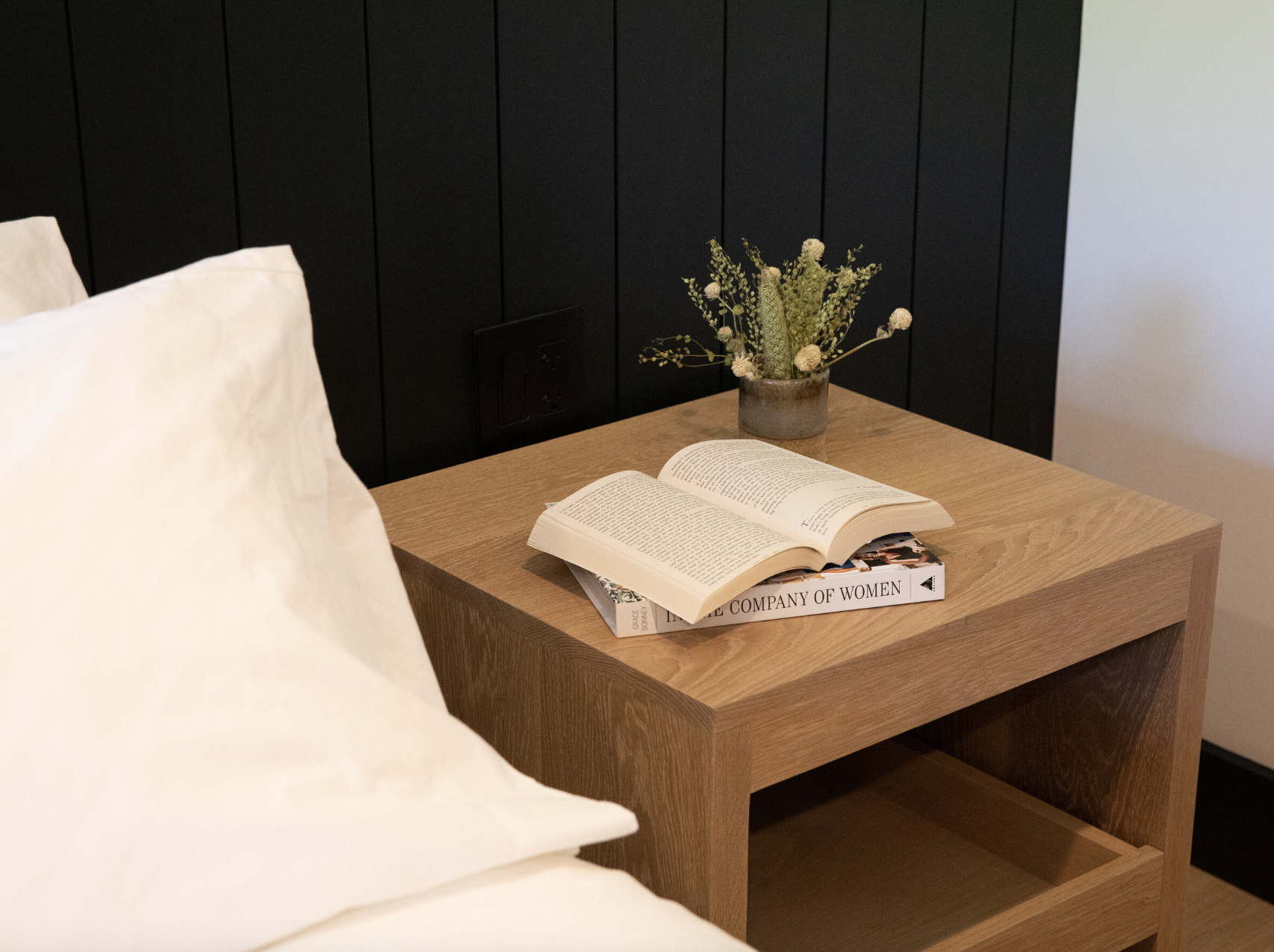 A bedside wooden table with an open book, a magazine titled "The Company of Women," and a vase with white and green flowers, next to a bed with white pillows, against a black paneled wall.