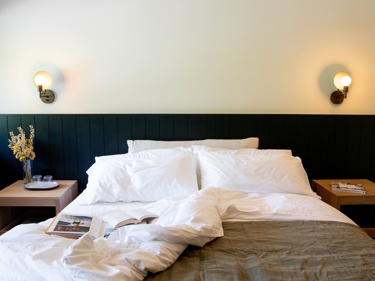 Unmade bed with white sheets, pillows, and a beige blanket, two nightstands with books and a vase of flowers, and wall-mounted lights in a modern bedroom.