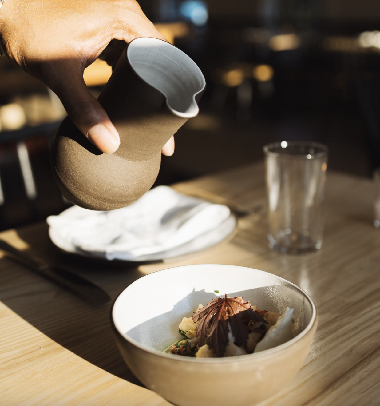 A hand pouring sauce over a plated dish of food in a white bowl on a wooden table in a restaurant.