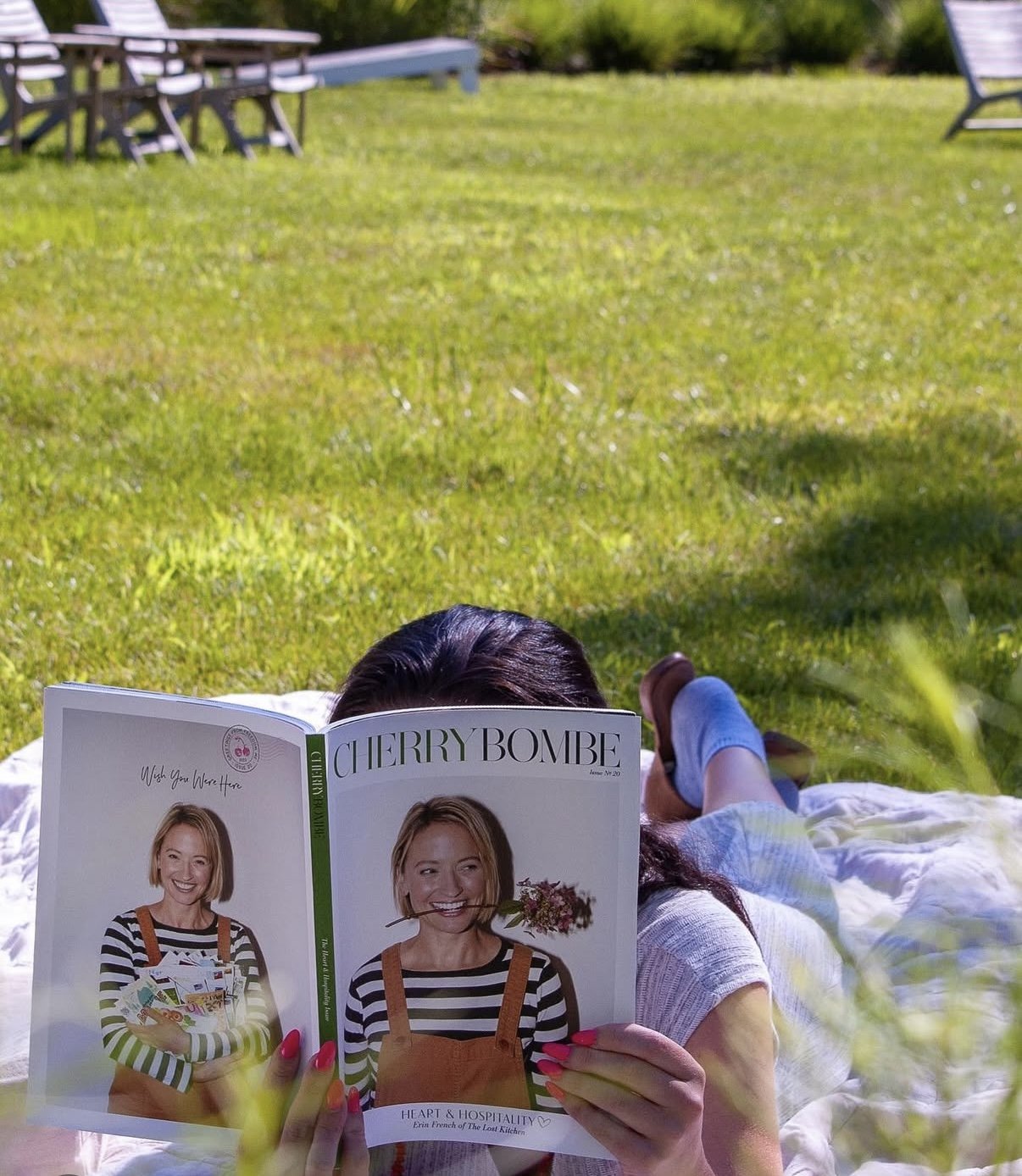 A person with dark hair lies on a blanket on the grass, reading a magazine titled 'Cherry Bombe'. The magazine's cover features a smiling woman with short blonde hair holding a bouquet of flowers in her mouth, dressed in striped shirt and overalls. T