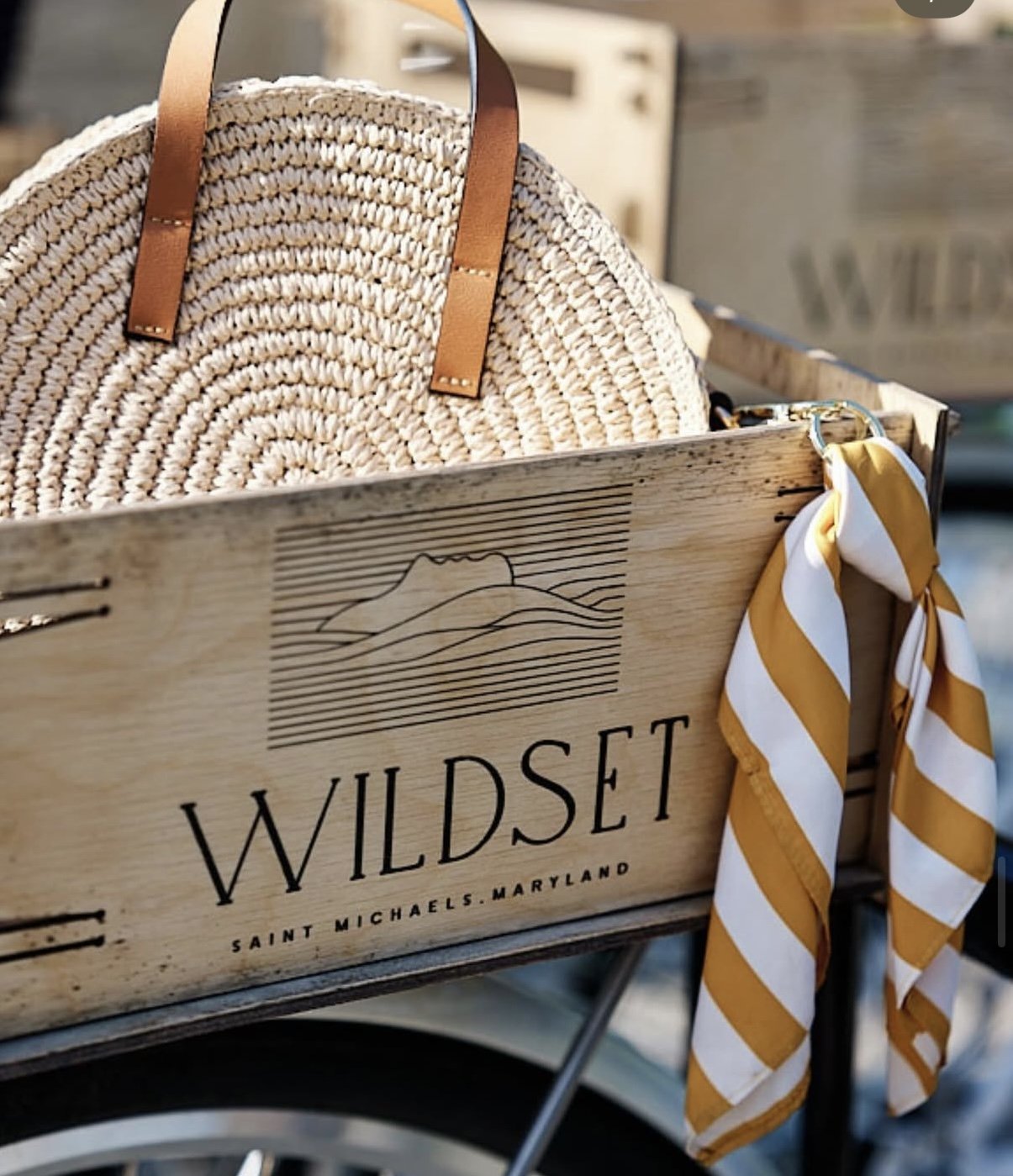 A wooden crate labeled 'Wildseed Saint Michaels Maryland' contains a woven straw bag with leather handles and a yellow and white striped scarf tied to the side.