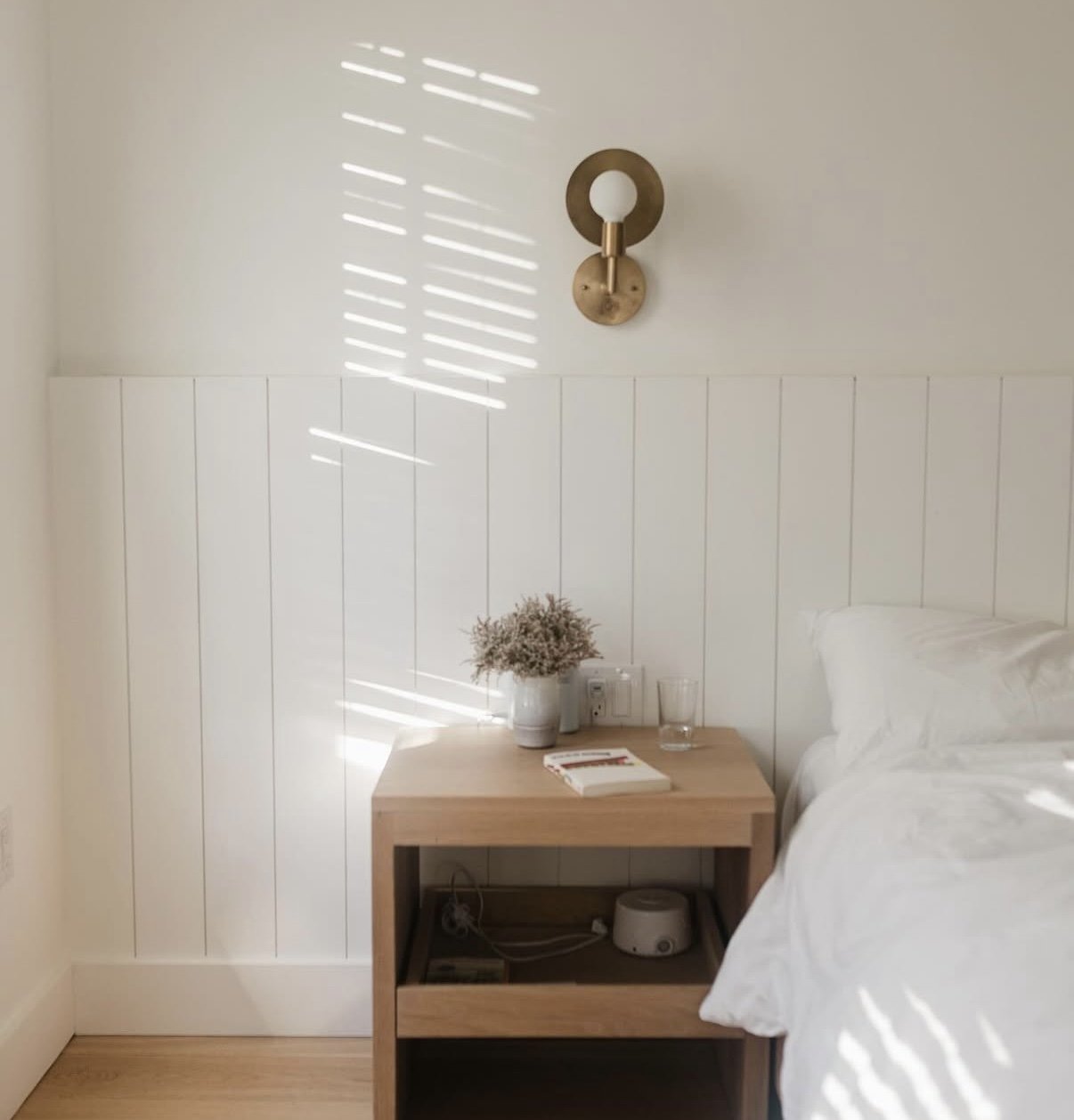 A bedroom corner with a wooden nightstand, a white flower vase, a book, a glass of water, a white bed, and a wall-mounted lamp. Sunlight creates horizontal stripe shadows on the wall.
