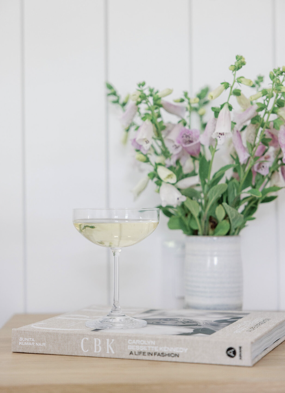 A glass of white wine on top of a magazine with a bouquet of pink and white flowers in the background, set on a wooden surface.