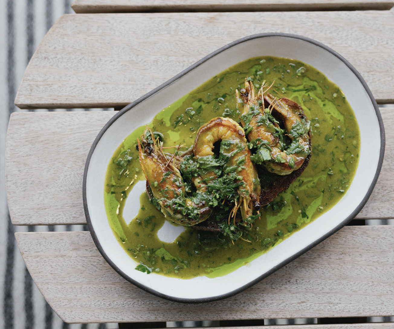 Shrimp atop a piece of toasted bread, garnished with fresh herbs, served with a vibrant green sauce.