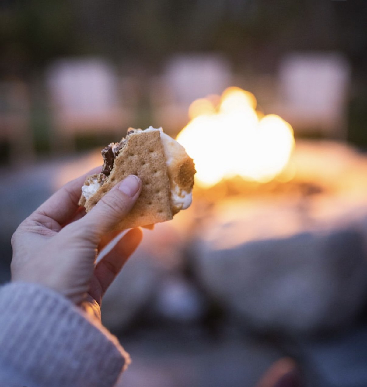 Person holding a s'mores treat with marshmallow and chocolate over a campfire at dusk.