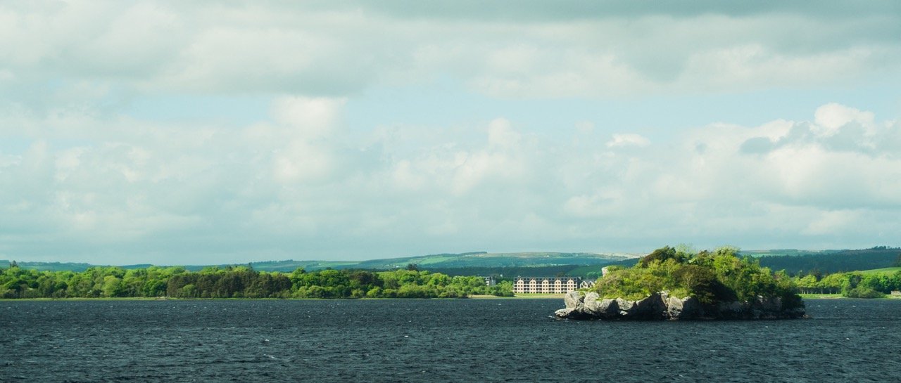 Escenario de un lago con una pequeña isla verde y rocas, fondo de colinas y un edificio grande, cielo nublado.
