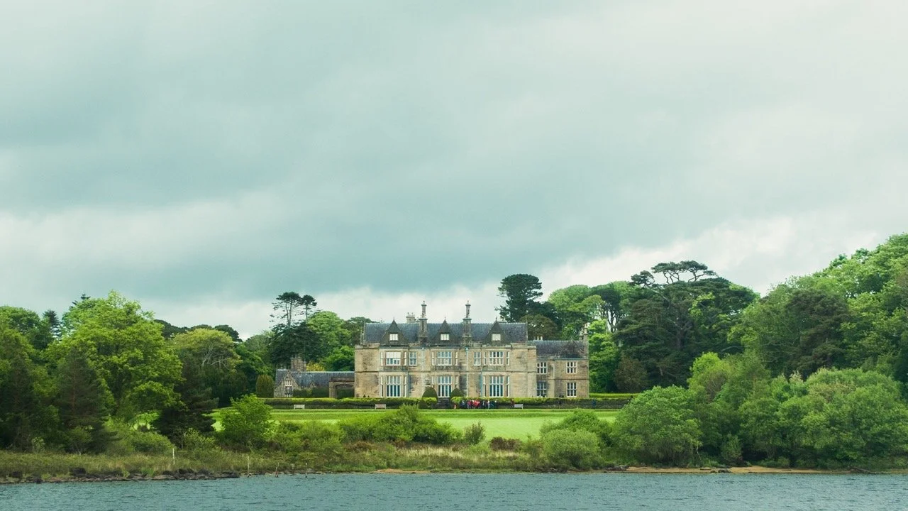 Palacio antiguo rodeado de árboles y jardines, con un lago en primer plano y un cielo nublado.