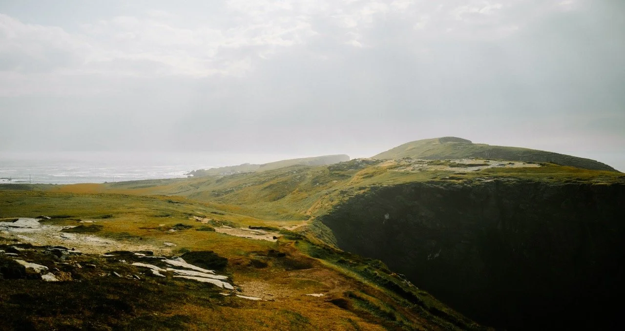 Paisaje de colinas verdes y acantilados, con cielo nublado y área costera en la distancia.