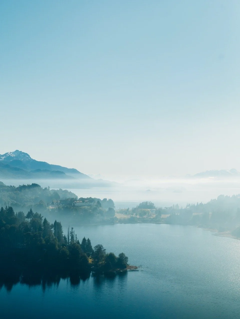 Paisaje de un lago rodeado de árboles y montañas con neblina y cielo despejado