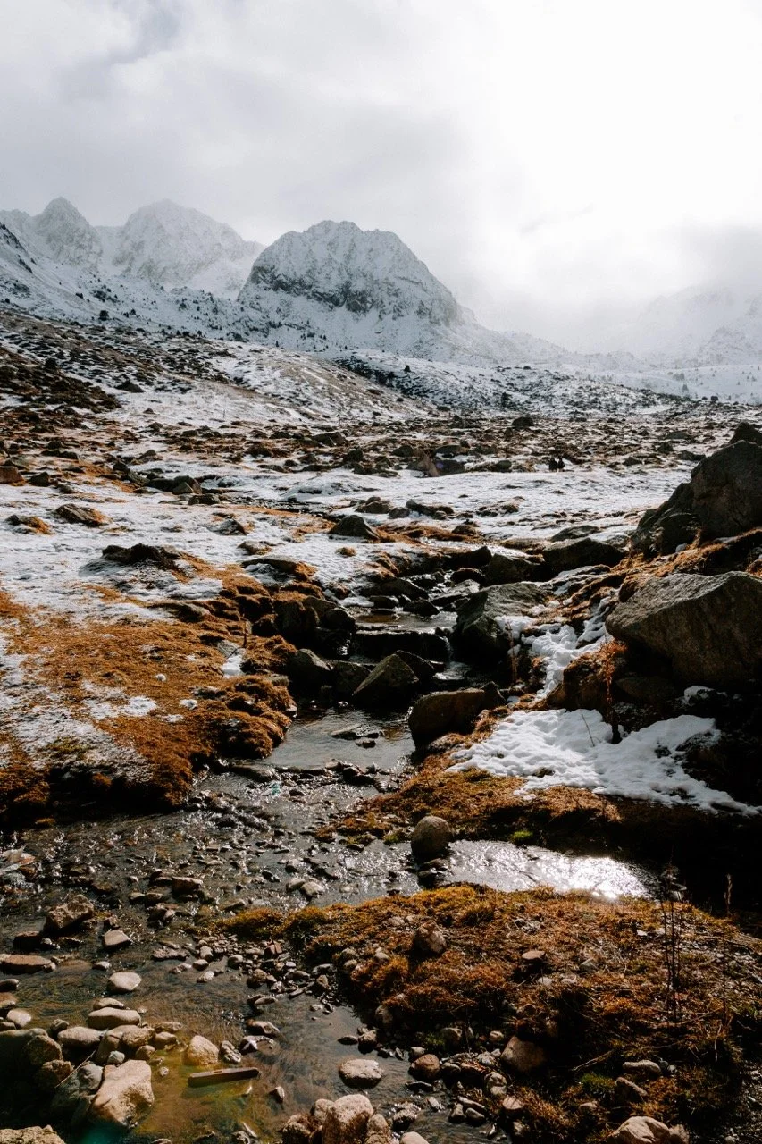 Paisaje montañoso con nieve, roca y río en primer plano, con montañas y nubes en el fondo