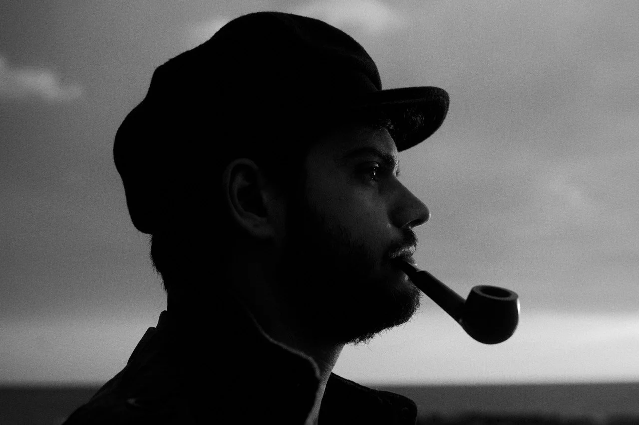 Perfil de un hombre con gorro y barba, fumando una pipa, en un entorno oscuro con cielo nublado.
