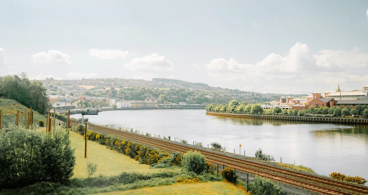 Vista panorámica de un río con vegetación y edificios al fondo, con vías del tren a la orilla.