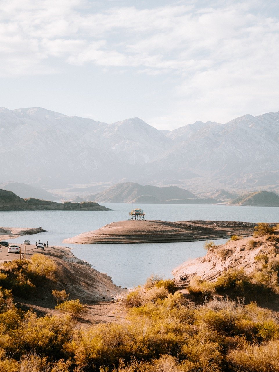 Paisaje de un lago rodeado de montañas con una estructura en el agua y vegetación en primer plano.