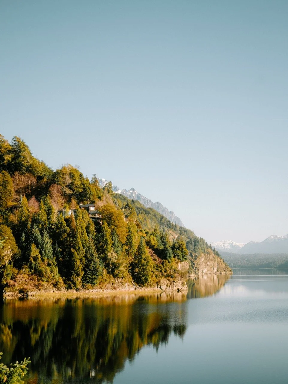 Paisaje de un lago rodeado de montañas y bosques verdes bajo un cielo claro.