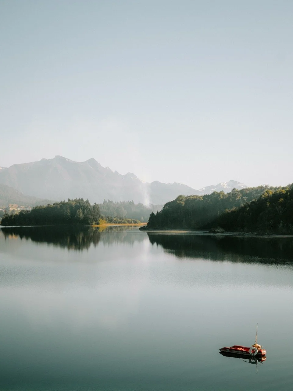 Un lago con agua tranquila, rodeado de colinas y montañas al fondo, y un bote rojo en el agua en la parte inferior derecha.