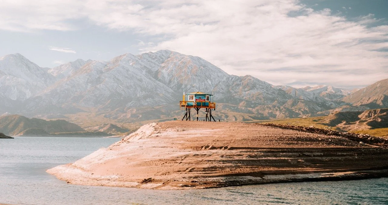 Una pequeña estructura de madera en un islote rodeado por agua, con montañas en el fondo y cielo parcialmente nublado.