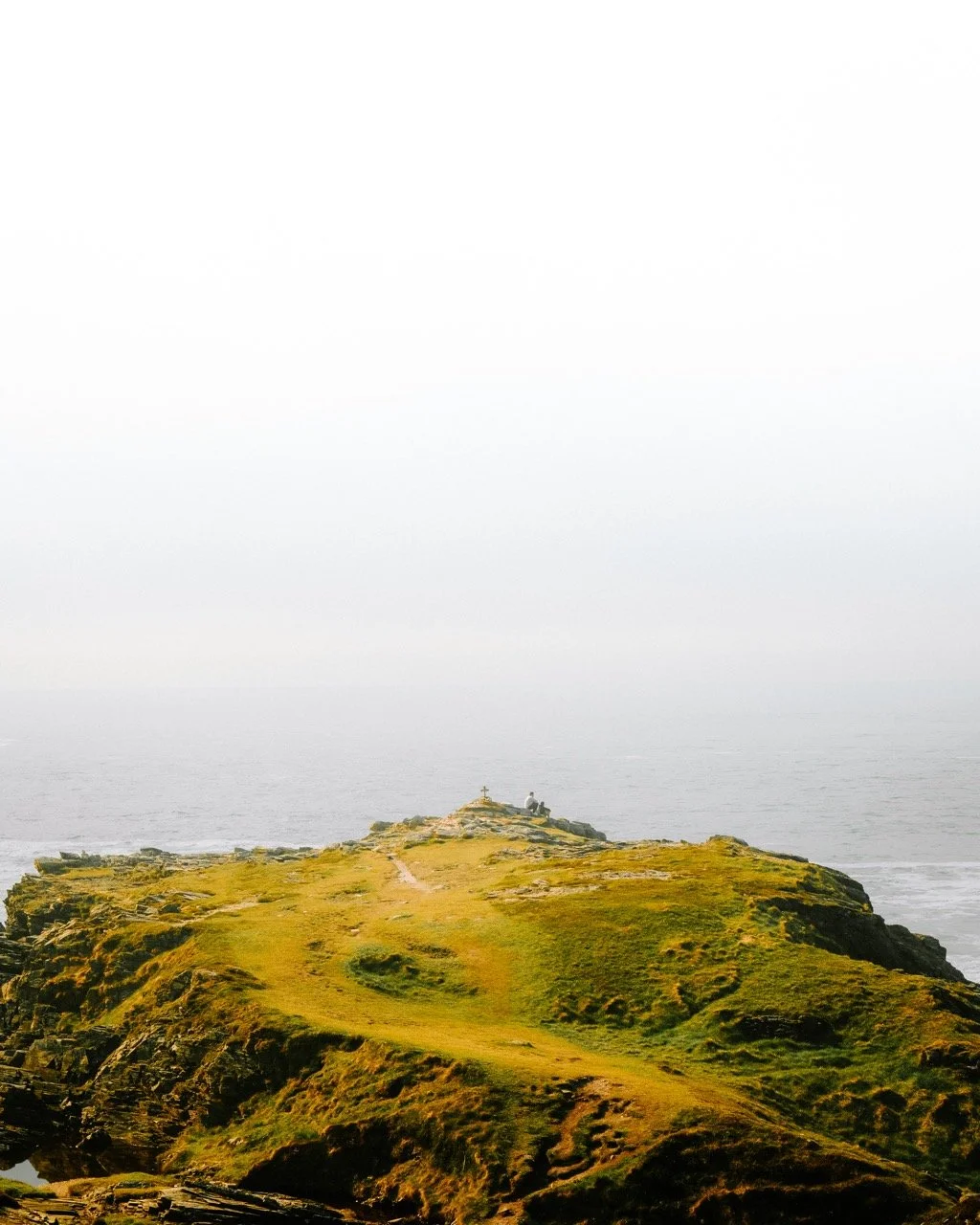 Cabo con césped y rocas, con cruz en la cima y vista al mar