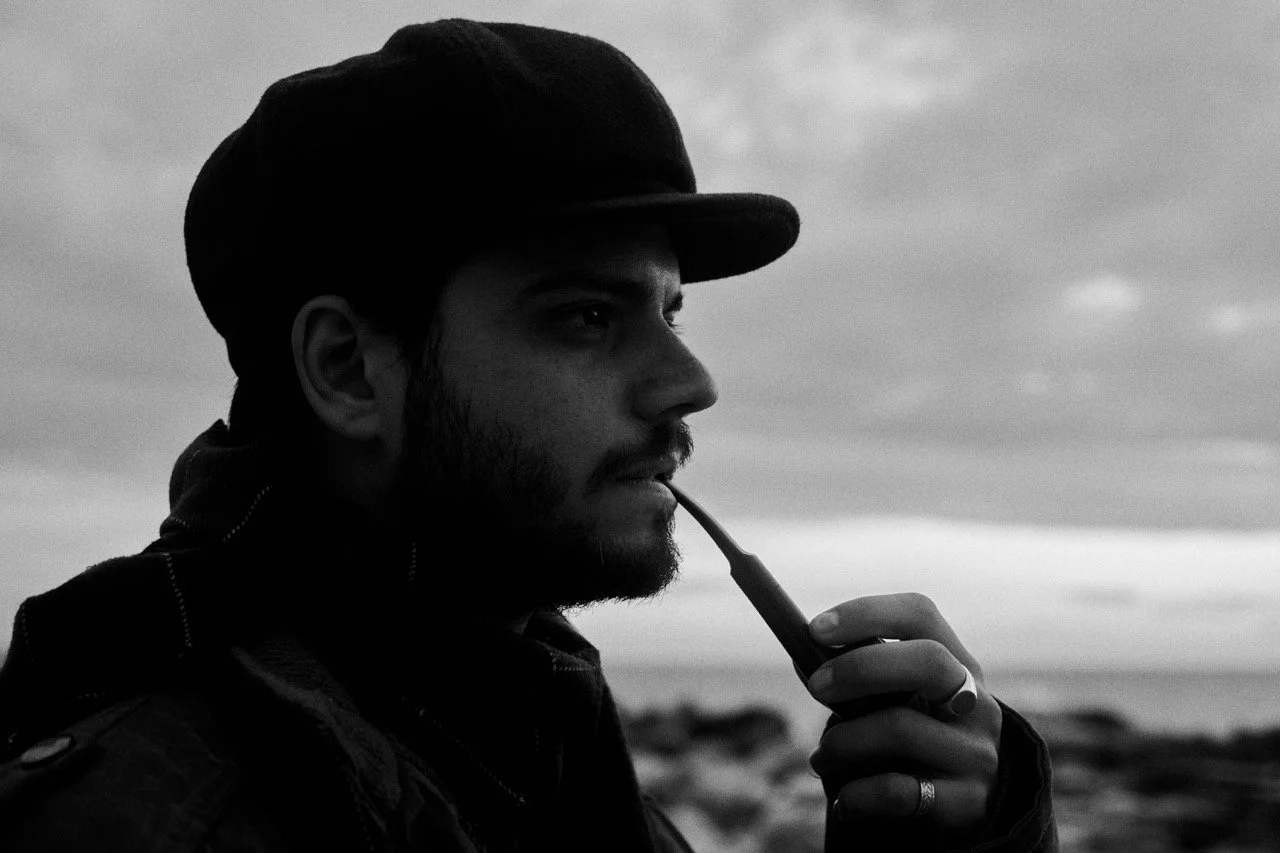 Joven hombre con barba y gorra negra, sosteniendo una pipa, mirando hacia la distancia en un paisaje con cielo nublado en blanco y negro.